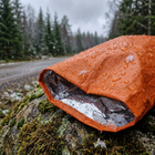 Regndråper og våt snø perler seg på den oransje nødsovsekken ved en grusvei, silversiden vises i åpningen