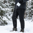 Vann helles fra en emaljekopp på sorte vinterbukser og perler seg og renner av stoffet i snøen