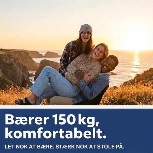 Tre grinende venner sidder sammen i campingstole på en klippe ved havet i solnedgang, med tekst om, at stolen bærer 150 kg