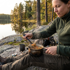 Kvinde spiser gryde fra en gryde med en sort multislev på en klippe ved en skovssø i aftenlys
