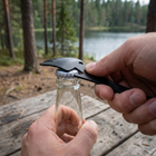 Multitool-flaskeåbner haget under kronen på en glasklasulle ved et campingbord i skoven